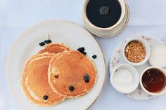 three pancakes with blueberries and syrup on a plate next to two cups of coffee