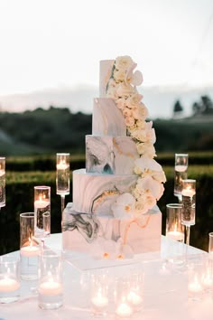 a wedding cake with white flowers and candles on the table in front of an outdoor setting