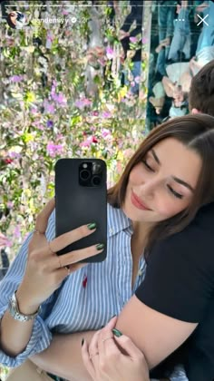 a woman holding a cell phone while sitting next to a man in front of flowers