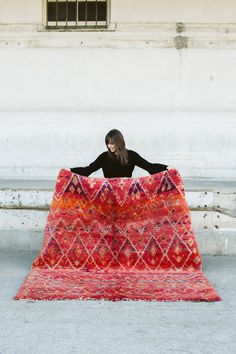 a woman sitting on top of a large red rug in front of a white building