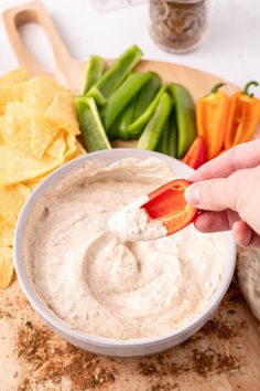 a person dipping dip into a white bowl with chips and vegetables around it on a cutting board