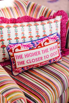 two colorful pillows sitting on top of a striped couch next to a pillow that says the higher the hair, the closer to god