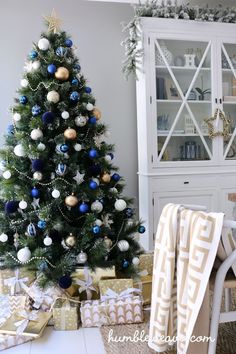 a christmas tree with blue and gold ornaments in front of a white hutch filled with presents