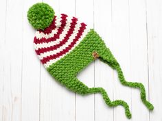 a knitted green frog hat with red and white stripes on the front, sitting on top of a white wooden surface