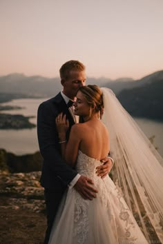 a bride and groom embracing each other on top of a mountain overlooking the lake at sunset