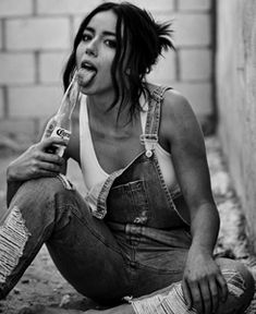 a woman sitting on the ground with a soda bottle in her mouth and wearing overalls