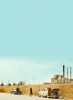 people walking down the side of a road next to parked cars and an industrial area