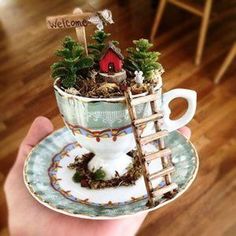 a cup filled with plants and a small bird house on top of it, held in someone's hand