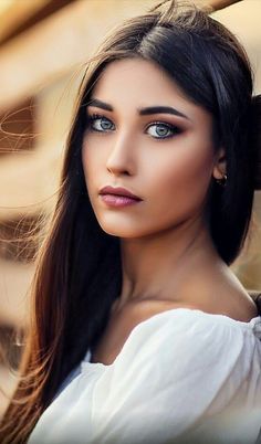 a woman with long hair leaning against a wooden wall and looking at the camera while wearing a white top