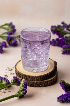 a glass filled with purple liquid sitting on top of a wooden coaster next to flowers