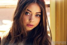 a beautiful young woman with long brown hair and blue eyes looking at the camera while standing in front of a window