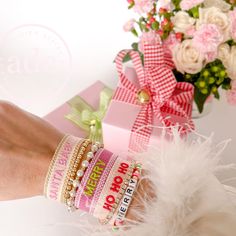 a woman's hand with bracelets and flowers in the background, next to a pink gift box