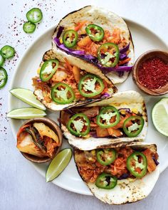 three tacos on a white plate with limes and red peppers next to them