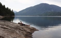 a man laying on top of a rock next to a body of water with mountains in the background