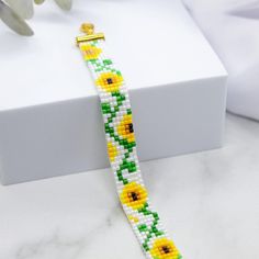 a yellow and green beaded bracelet sitting on top of a white box next to flowers