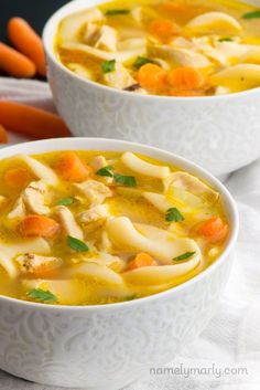 two white bowls filled with chicken noodle soup on top of a table next to carrots