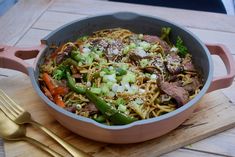 a pot filled with noodles and vegetables on top of a wooden cutting board next to a fork