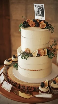 a three tiered white cake sitting on top of a wooden table next to cupcakes