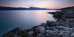 the sun is setting over the water and mountains in the distance, with rocks on either side