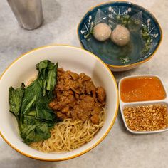 two bowls filled with noodles and meat next to dipping sauces on a marble table