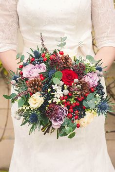 a woman in a white dress holding a red and purple bouquet with pine cones on it