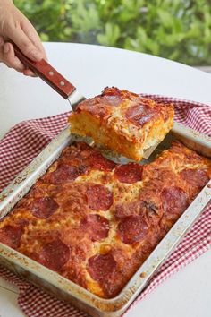 someone cutting into a pepperoni casserole with a pizza spatula on a red and white checkered tablecloth