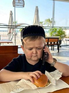 a young boy sitting at a table while talking on a cell phone and eating a hamburger