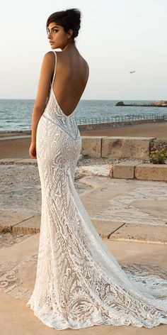 a woman in a white wedding dress standing on the beach looking at the ocean with her back to the camera