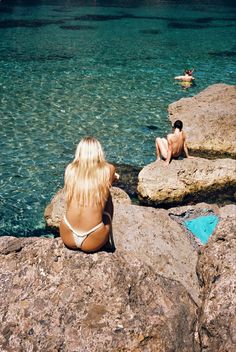 two people are sitting on rocks near the water and one person is swimming in the ocean