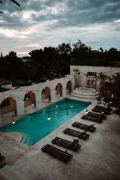 an outdoor swimming pool with lounge chairs and lights around it at dusk or dawn in the evening