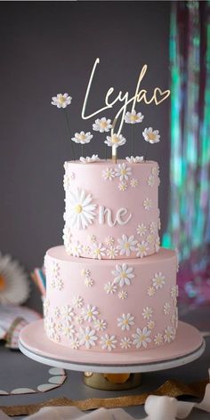 a pink cake with daisies on top and the word love spelled in gold letters