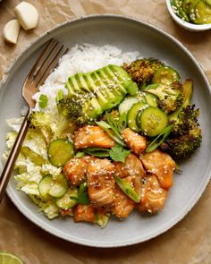 a white plate topped with chicken, broccoli and cucumbers next to rice