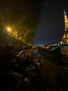 some people sitting on the side of a river at night with the eiffel tower in the background