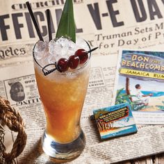 a tall glass filled with ice and cherries on top of a table next to newspapers