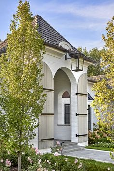 a white house with a black roof and some trees in the front yard, along with pink flowers