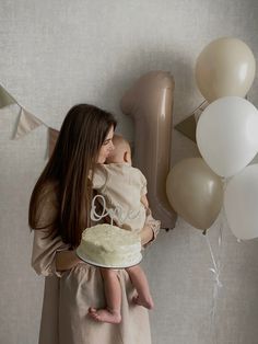 a woman holding a baby in her arms while she holds a cake with the number one on it