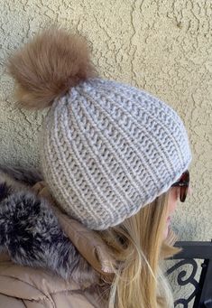 a woman with blonde hair wearing a white knitted beanie and fur pom - pom