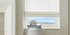 a white vase sitting on top of a table next to a window covered in blinds