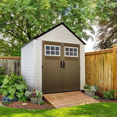 a small shed in the middle of a yard with flowers and plants around it, next to a wooden fence