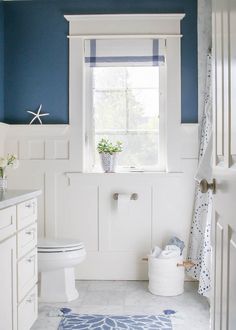 a white toilet sitting next to a window in a bathroom under a blue and white rug