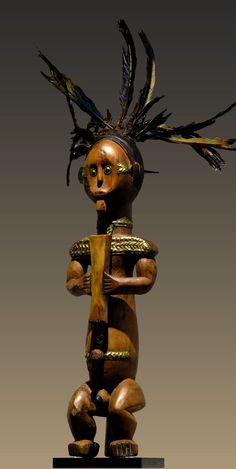 a wooden statue with feathers on it's head and arms, standing in front of a gray background