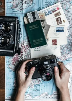 a person holding a camera on top of a map next to some stamps and other items