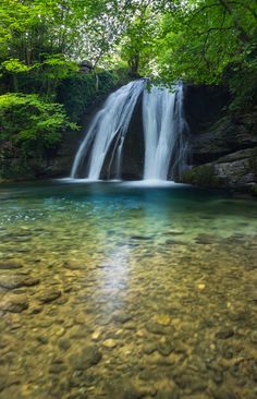 a waterfall in the middle of a forest filled with green trees and rocks, surrounded by clear water