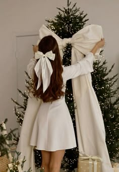 a woman standing in front of a christmas tree wearing a white dress and bow tie