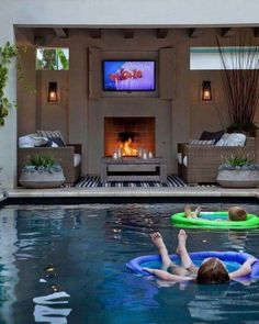 two people floating in a swimming pool with an open fire place and television on the wall