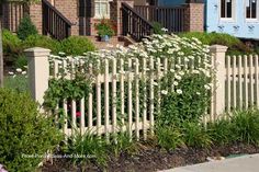 a white picket fence in front of a brick house with flowers growing on the side