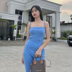 a woman in a blue jumpsuit is holding a brown purse and posing for the camera