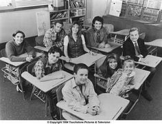 black and white photograph of students in classroom with teacher sitting at desks smiling for the camera