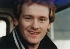 a close up of a person wearing a scarf and smiling at the camera with a bus in the background