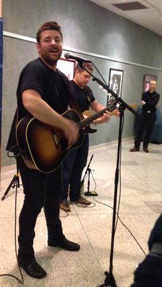 a man holding a guitar while standing in front of a microphone and another man playing the guitar